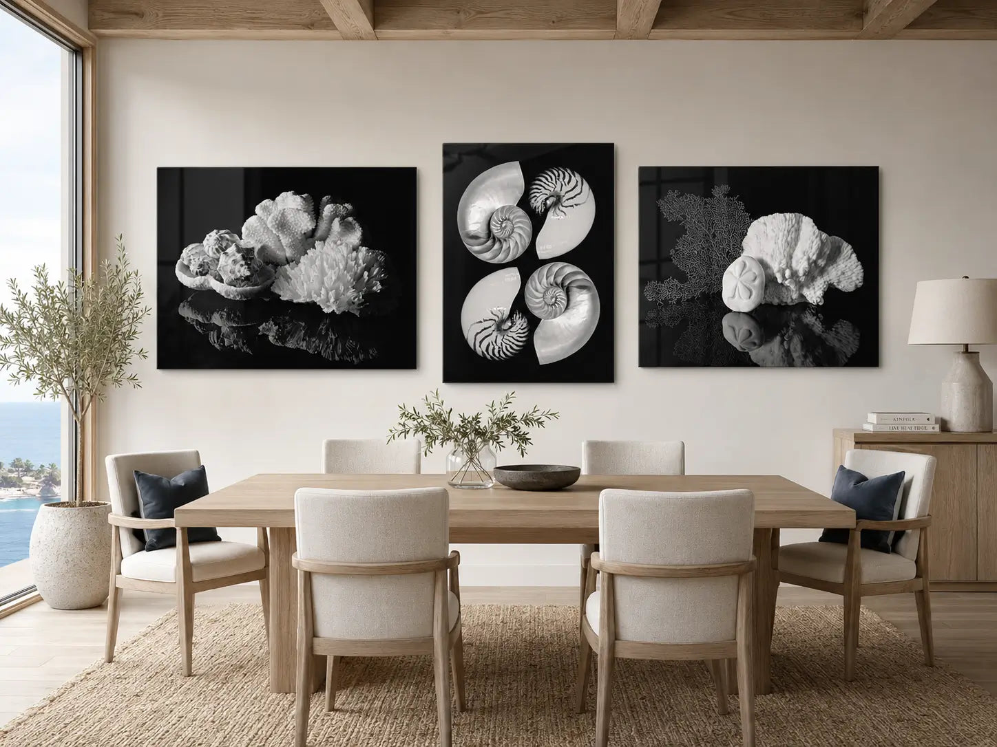 Three black-and-white shell and coral glass prints displayed above a warm neutral coastal dining table with slate blue accents and an ocean view.