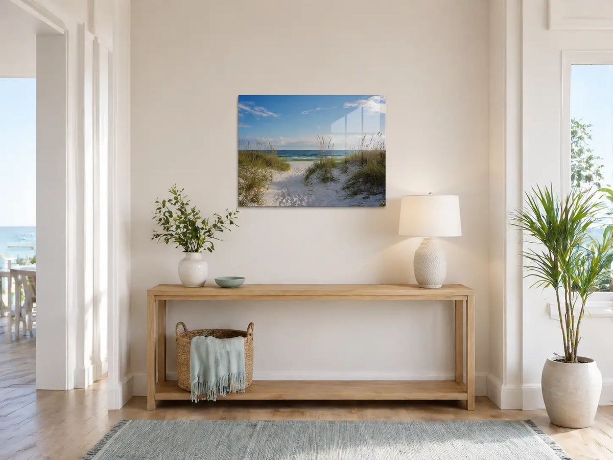 Bright coastal entryway with a frameless glass print of a sandy dune path leading to the beach above a light wood console table.