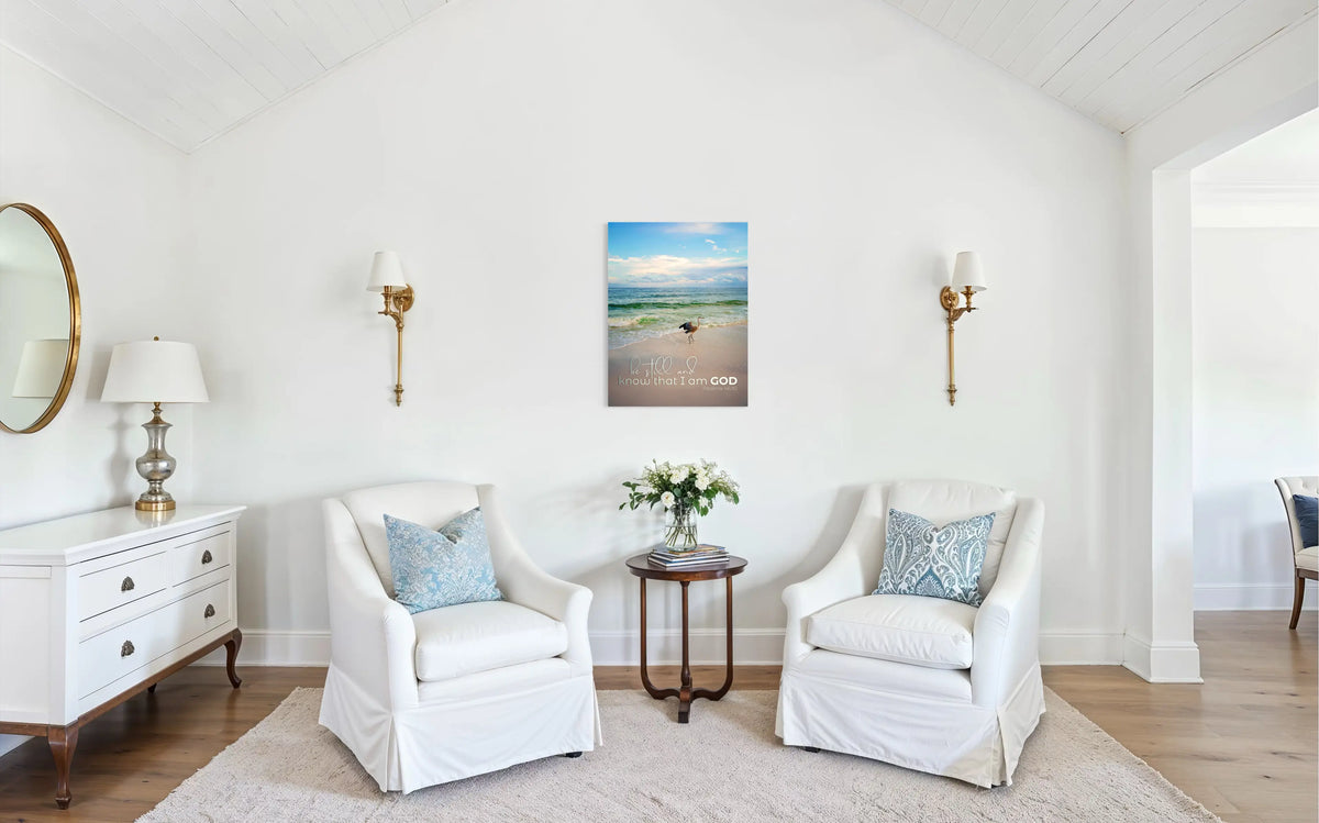 Coastal Grandma sitting room with white slipcovered armchairs, blue patterned pillows, and beach wall art that reads “Be still and know that I am God.”