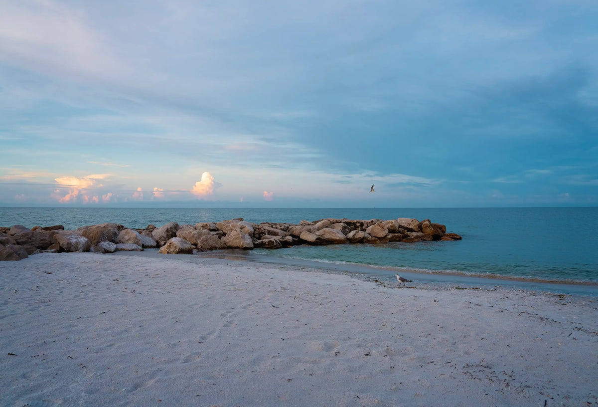 Quiet shoreline at St. Pete’s Beach with pale blue water, soft clouds, a low rock line, and a single gull.