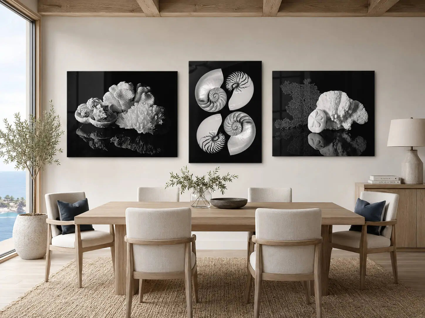 Three black-and-white shell and coral glass prints displayed above a warm neutral coastal dining table with slate blue accents and an ocean view.