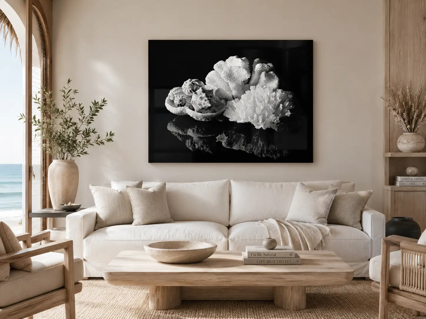 Black-and-white coral and seashell glass wall art displayed above a cream sofa in a warm modern coastal living room with an ocean view.